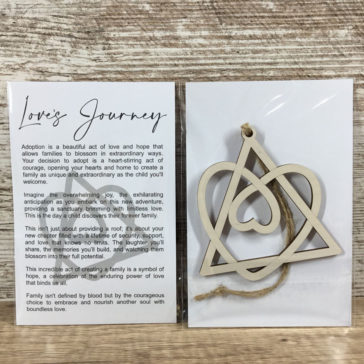 A wooden heart-shaped pendant with a heart outline and a small hanging loop, displayed next to a printed card titled 'Love's Journey' on a light wood surface.