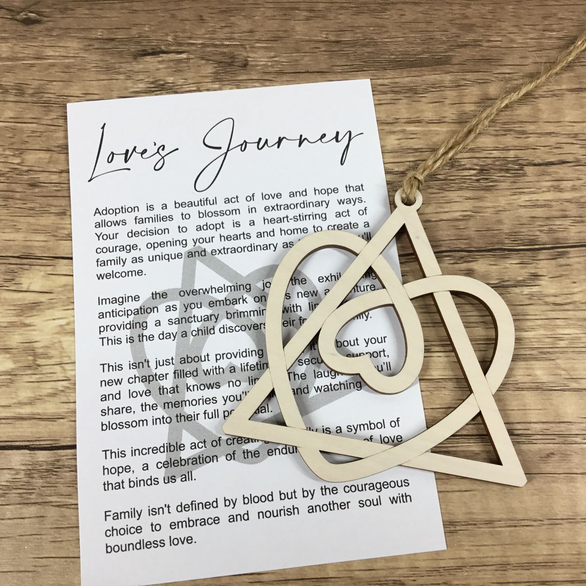 A wooden cutout ornament resting on a printed card titled 'Love's Journey' on a rustic wooden surface. The ornament is made of light natural wood and hangs from a piece of twine. 