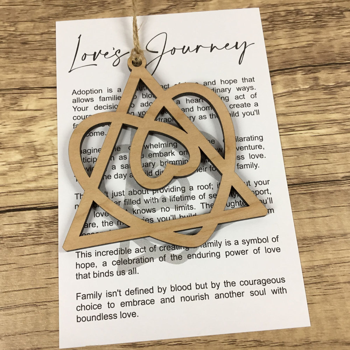 A light natural wood ornament shaped like a triangle enclosing a heart, hanging from a string over a printed card on a wooden surface.