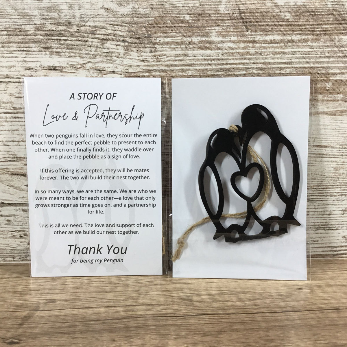 A penguin keepsake gift set on a light wood background, featuring a printed card that reads 'A Story of Love & Partnership' and a small black penguin wood figurine with a twine hanger.