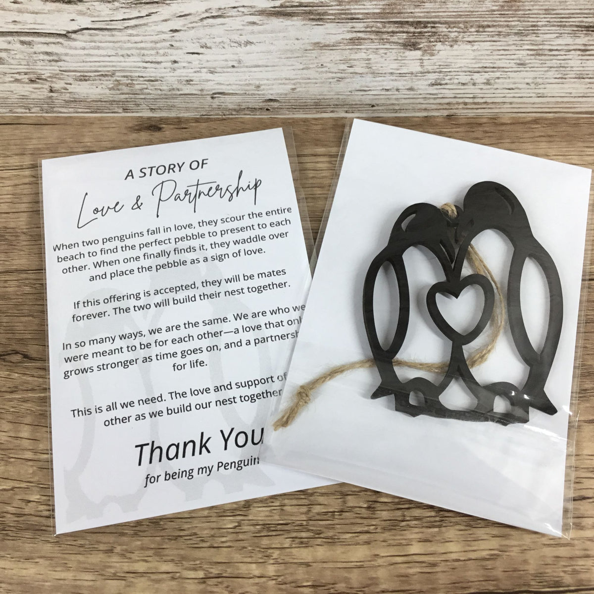 A black wood penguin nesting heart ornament on a white card with a small tag, displayed next to a printed 'A Story of Love & Partnership' thank you card on a light wood background.