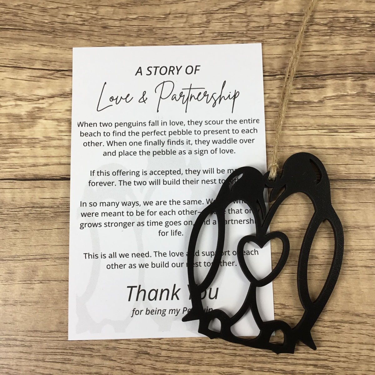 A printed card with penguin-themed wedding or partnership text lies on a wooden surface, with a black wood penguin cutout ornaments placed on top.