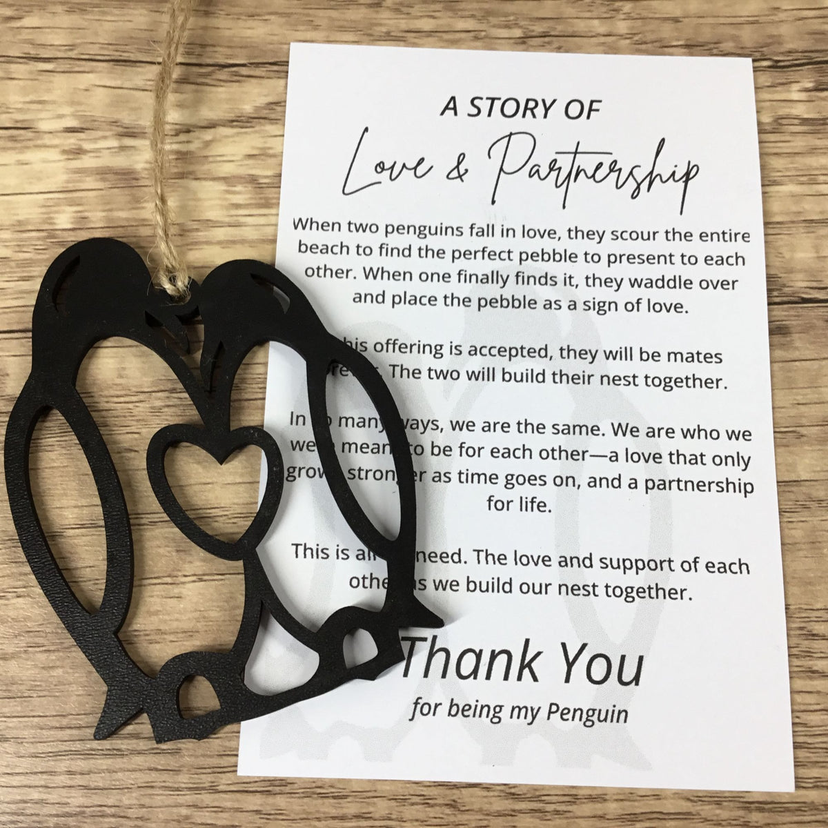A black laser-cut penguin nesting gift set on a light wood background, featuring a penguin silhouette with nested heart outlines and a printed penguin-themed thank you card.