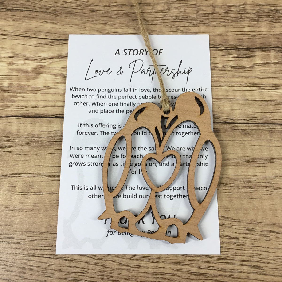 A wooden penguin-shaped ornament with a heart-shaped center cutout hangs from a twine string over a printed card on a wood surface.