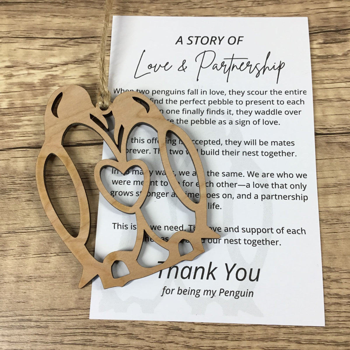 A wooden penguin-shaped ornament lies on top of a printed card that reads 'A STORY OF Love & Partnership' and 'thank You for being my Penguin,' placed on a wood surface.