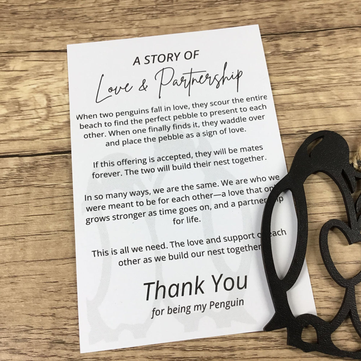A printed card reading 'A STORY OF Love & Partnership' with a poem about penguins and 'Thank You for being my Penguin' at the bottom, placed on a wooden surface next to a black wood penguin ornament.
