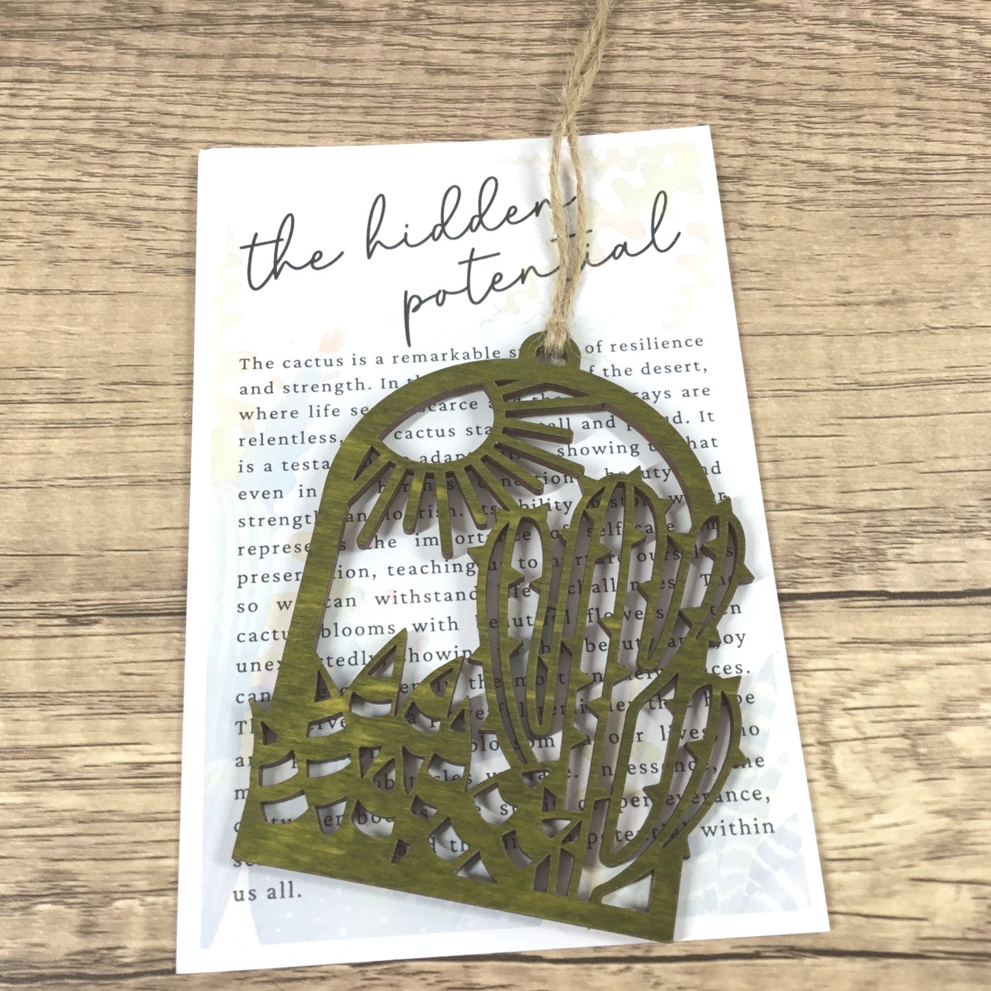 A laser-cut olive green wood cactus ornament with a sunburst pattern, hanging from a piece of twine over a printed card on a wooden surface.