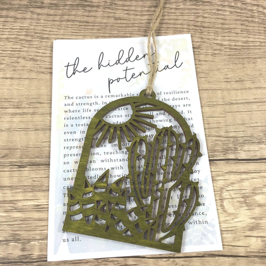 A laser-cut olive green wood cactus ornament with a sunburst pattern, hanging from a piece of twine over a printed card on a wooden surface.
