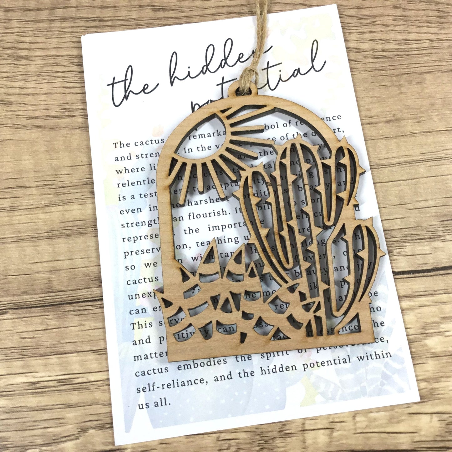 A laser-cut wooden ornament shaped like a cactus and sun, hanging from a string on a printed card over a wood surface.