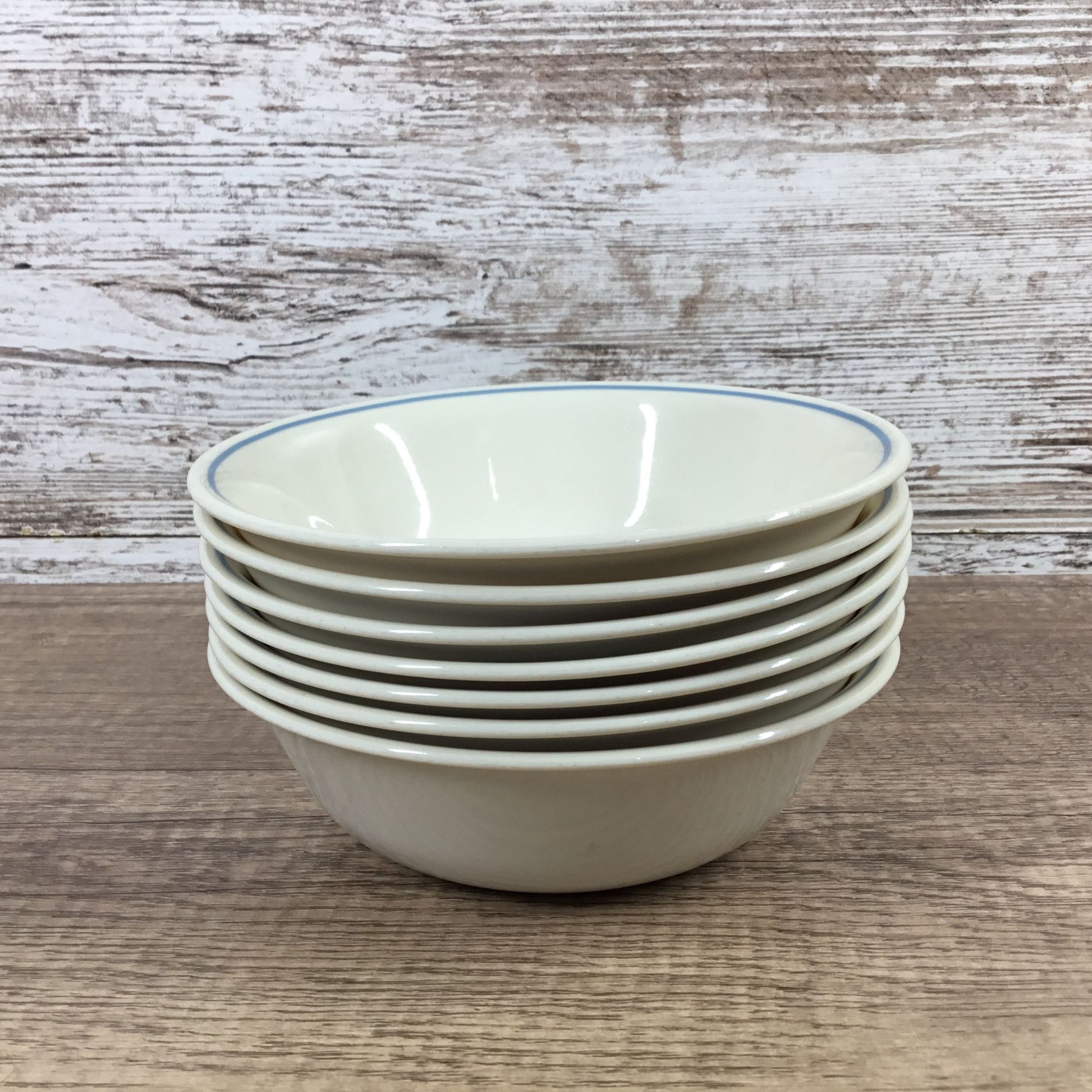 A stack of six Corelle bowls with a light beige/cream glaze and a thin blue rim, set on a rustic wooden surface against a distressed wood plank background.