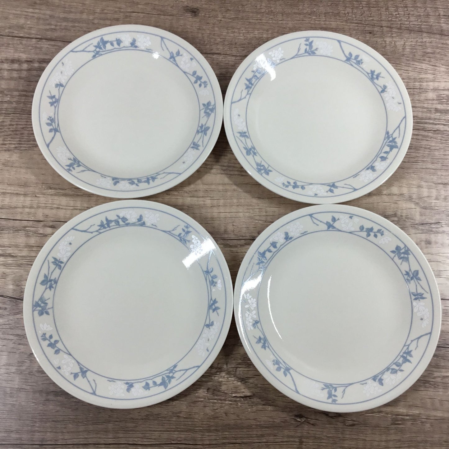 Four white Corelle plates with a blue and white floral border pattern, arranged on a wooden surface.