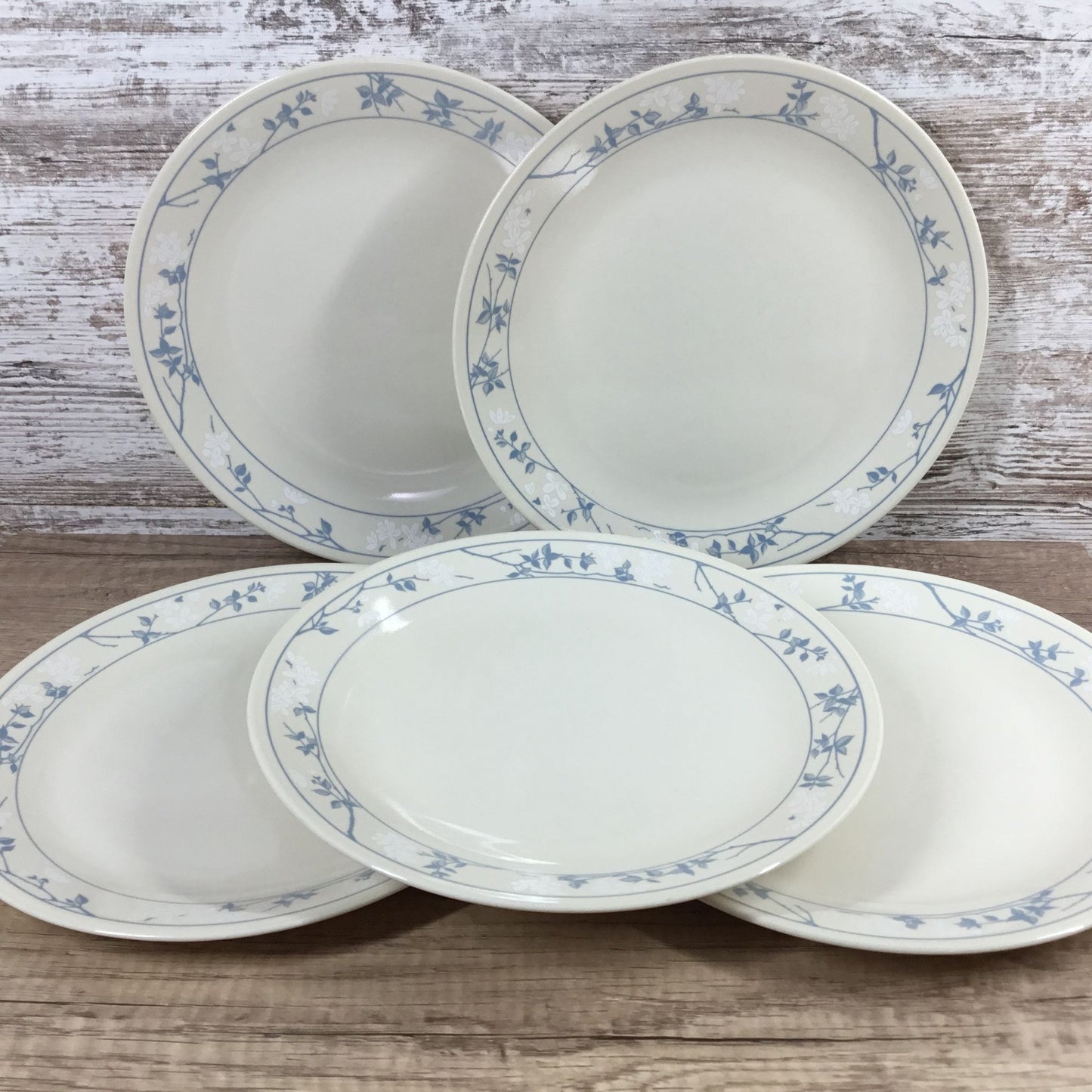 Five white Corelle plates with a delicate blue floral vine border, arranged on a rustic wooden surface against a whitewashed wood plank background.
