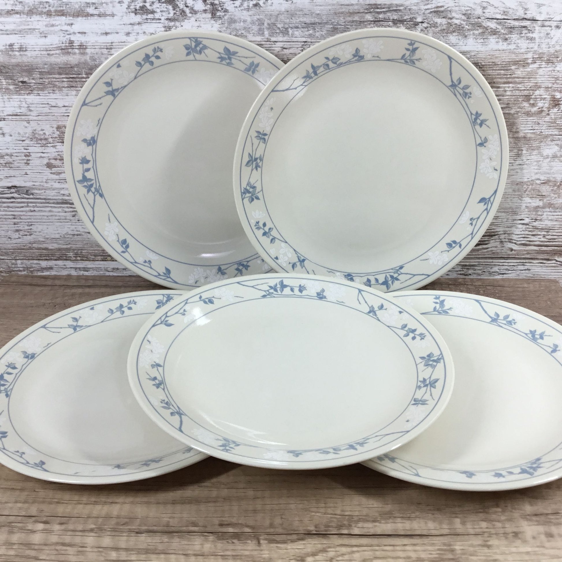 Five white Corelle plates with a delicate blue floral vine border, arranged on a rustic wooden surface against a whitewashed wood plank background.