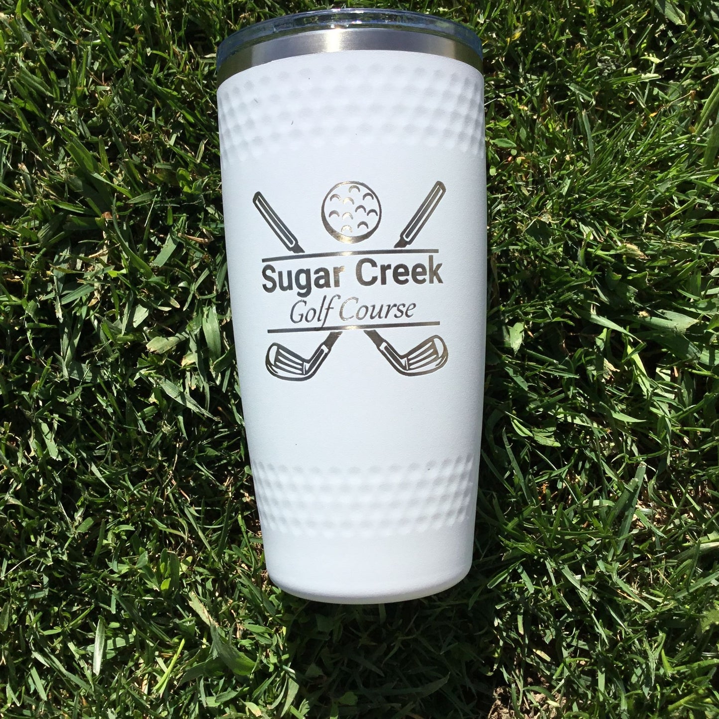A white 20 oz stainless steel tumbler with a dimpled golf ball design, featuring the text 'Sugar Creek Golf Course' and golf clubs graphics, placed on a grass surface.