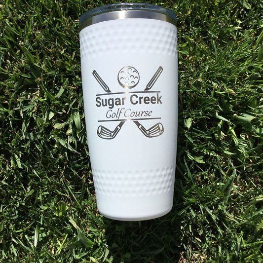 A white 20 oz stainless steel tumbler with a dimpled golf ball design, featuring the text 'Sugar Creek Golf Course' and golf clubs graphics, placed on a grass surface.