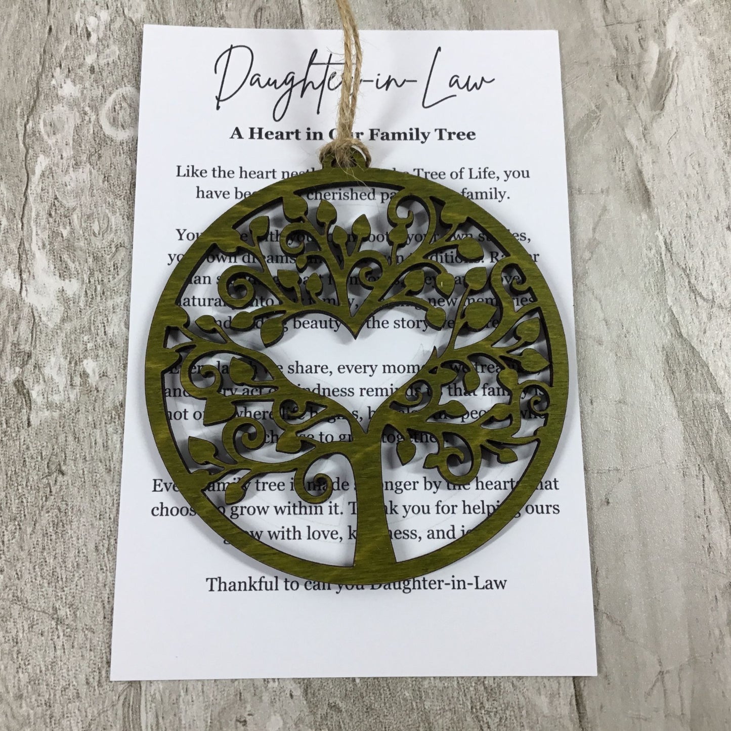 An olive green laser-cut tree of life ornament with a hanging loop, displayed on top of a white printed card that reads 'Daughter-in-Law: A Heart in Our Family Tree,' set against a light gray wood background.