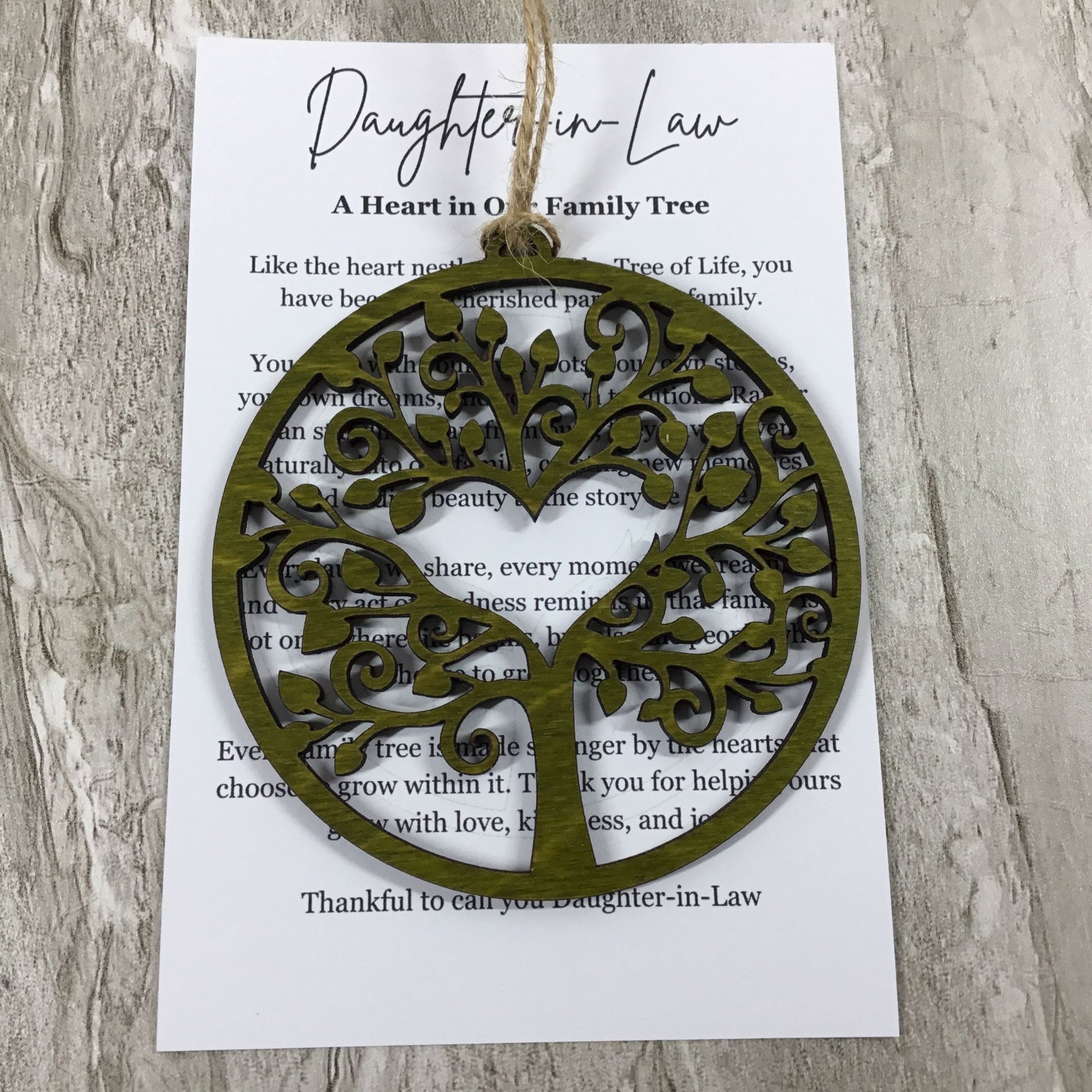 An olive green laser-cut tree of life ornament hangs from a printed card that reads 'Daughter-in-Law A Heart in Our Family Tree' on a light wood background.