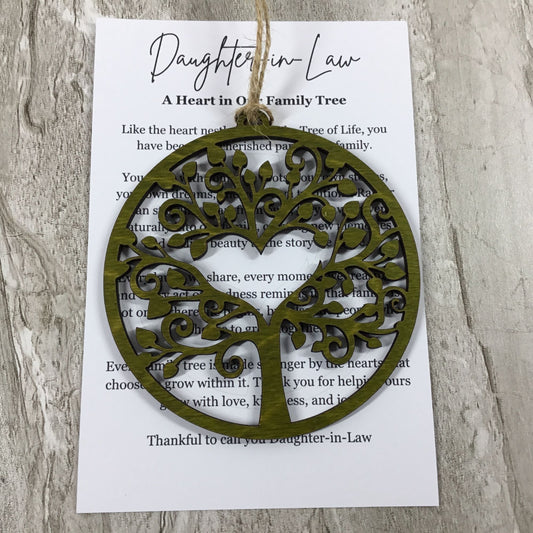 An olive green laser-cut tree of life ornament hangs from a printed card that reads 'Daughter-in-Law A Heart in Our Family Tree' on a light wood background.