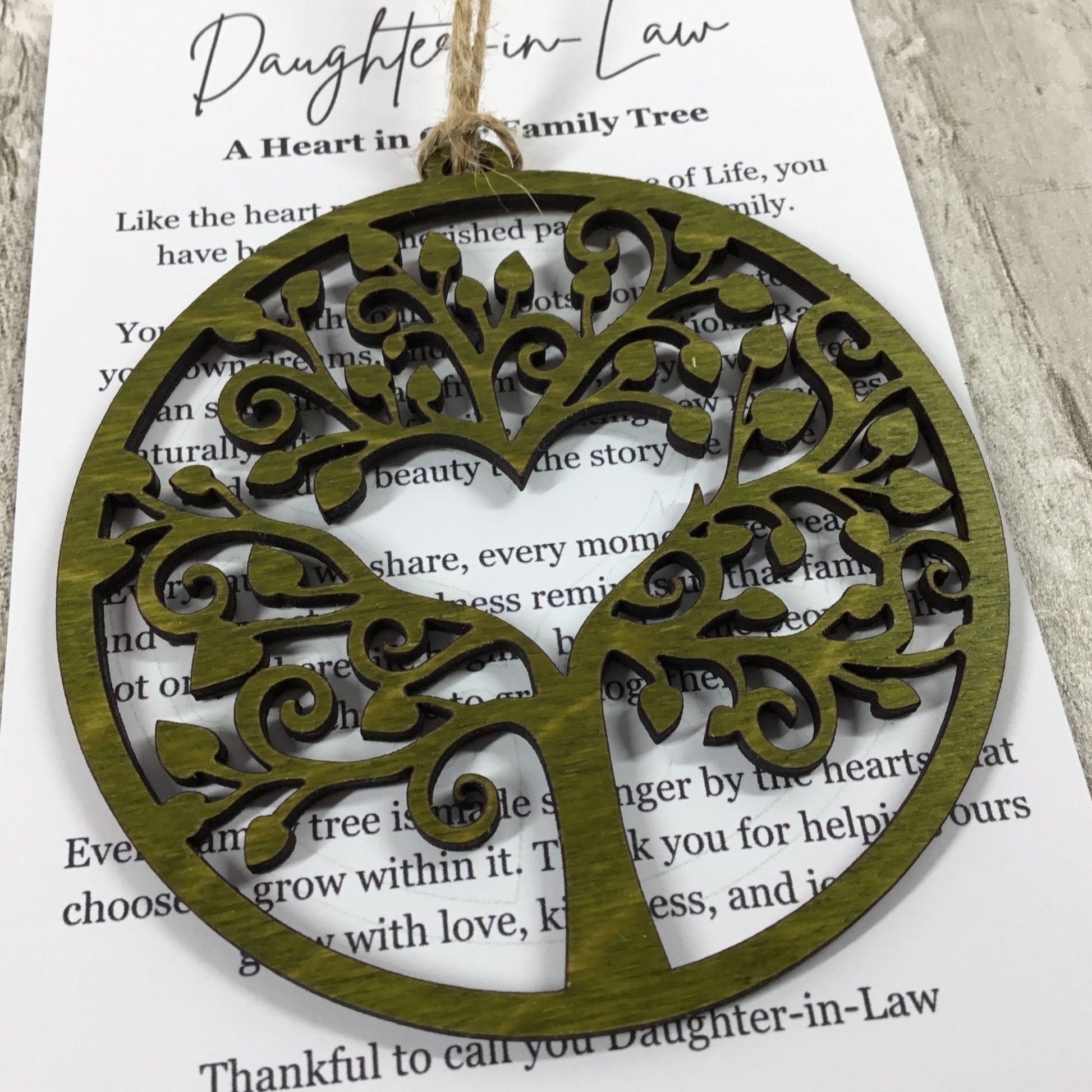 A round wooden ornament with a laser-cut tree of life design and a heart-shaped trunk, hanging from a twine string over a printed card that reads 'Daughter-in-Law'.