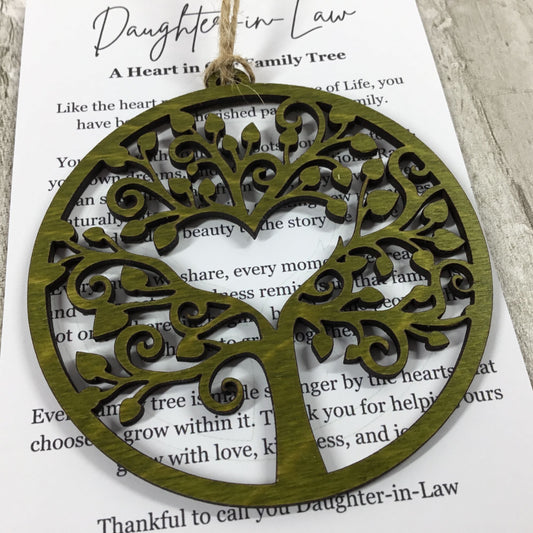 A round wooden ornament with a laser-cut tree of life design and a heart-shaped trunk, hanging from a twine string over a printed card that reads 'Daughter-in-Law'.