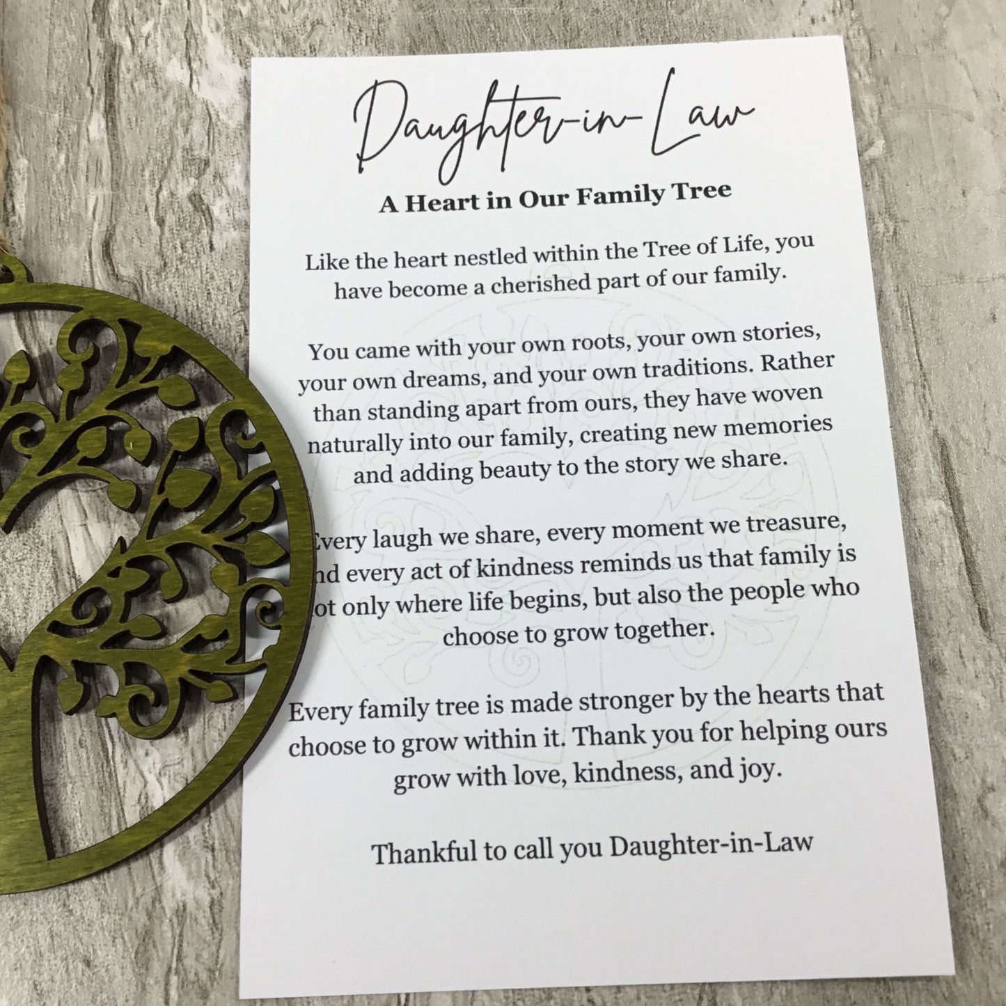 A printed card reading 'Daughter-in-Law: A Heart in Our Family Tree' with family tree text, styled on a light wood surface with a green decorative metal object partially visible at the left edge.