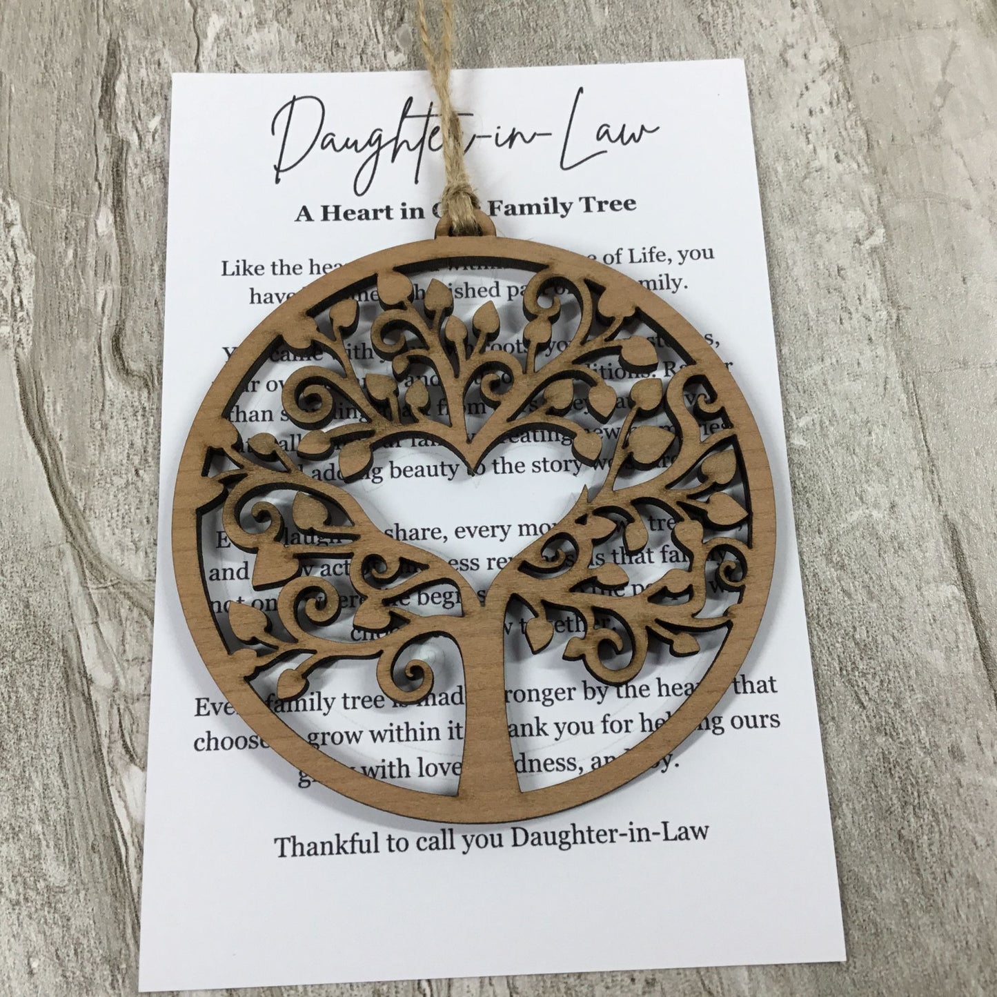 A round laser-cut wooden tree ornament hanging from a string over a printed card addressed 'Daughter-in-Law' on a light wood surface.