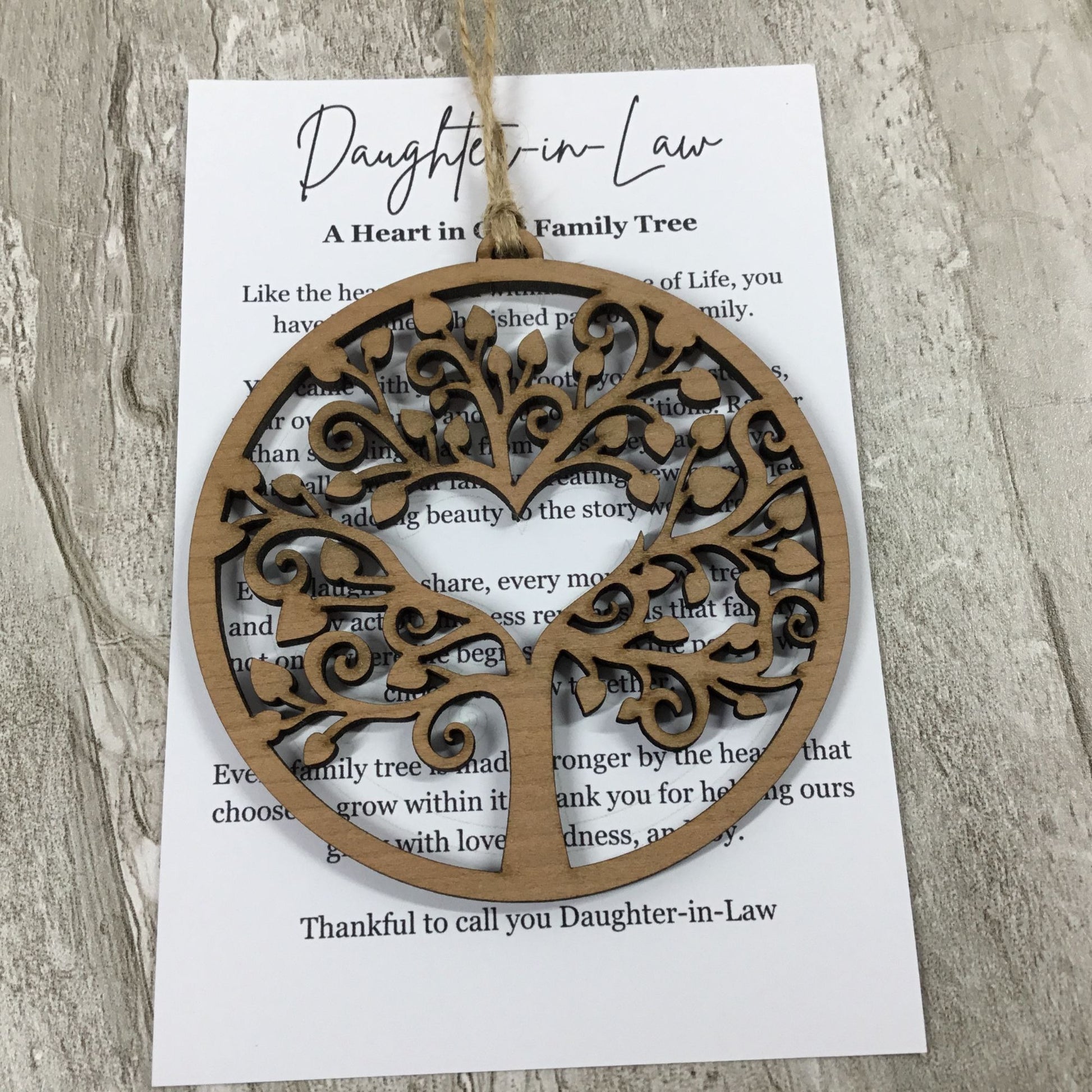 A round laser-cut wooden tree ornament hanging from a string over a printed card addressed 'Daughter-in-Law' on a light wood surface.