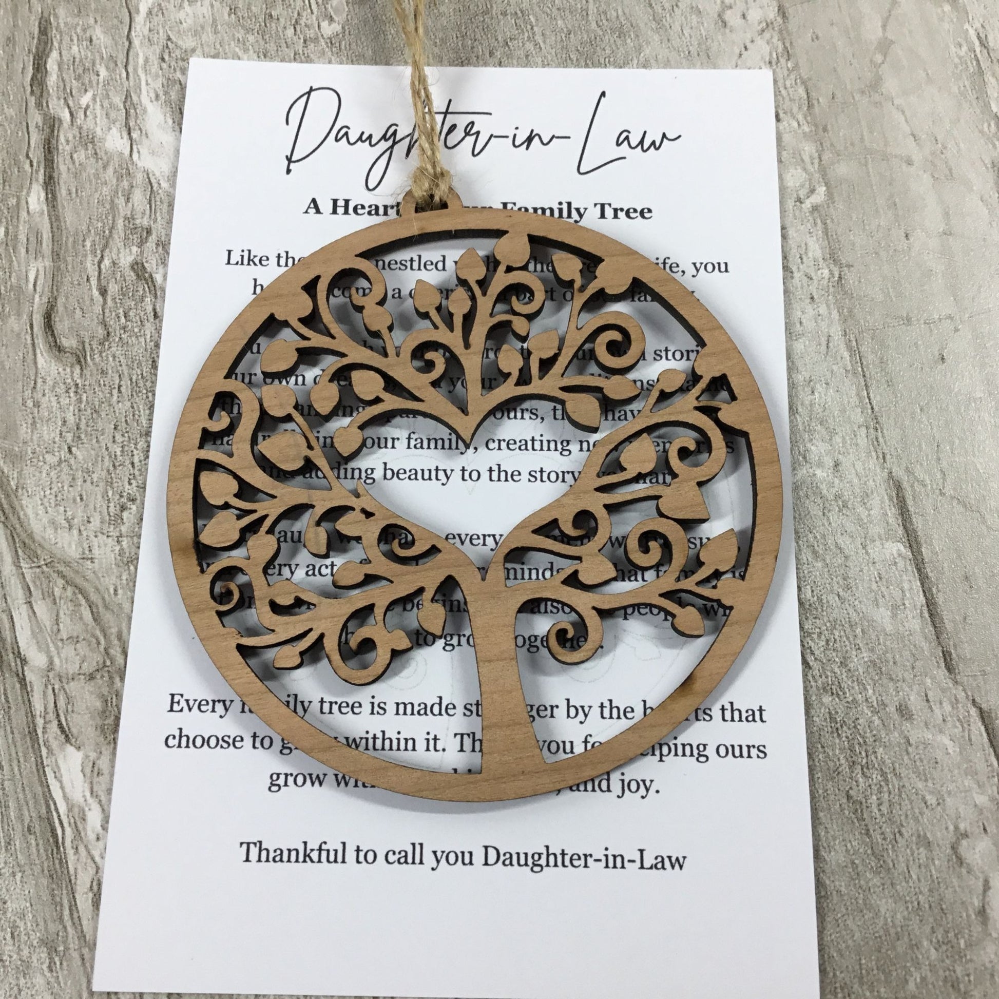 A round laser-cut wooden tree ornament hanging from a string, displayed over a printed card that reads 'Daughter-in-Law A Heart Family Tree' on a light wood surface.