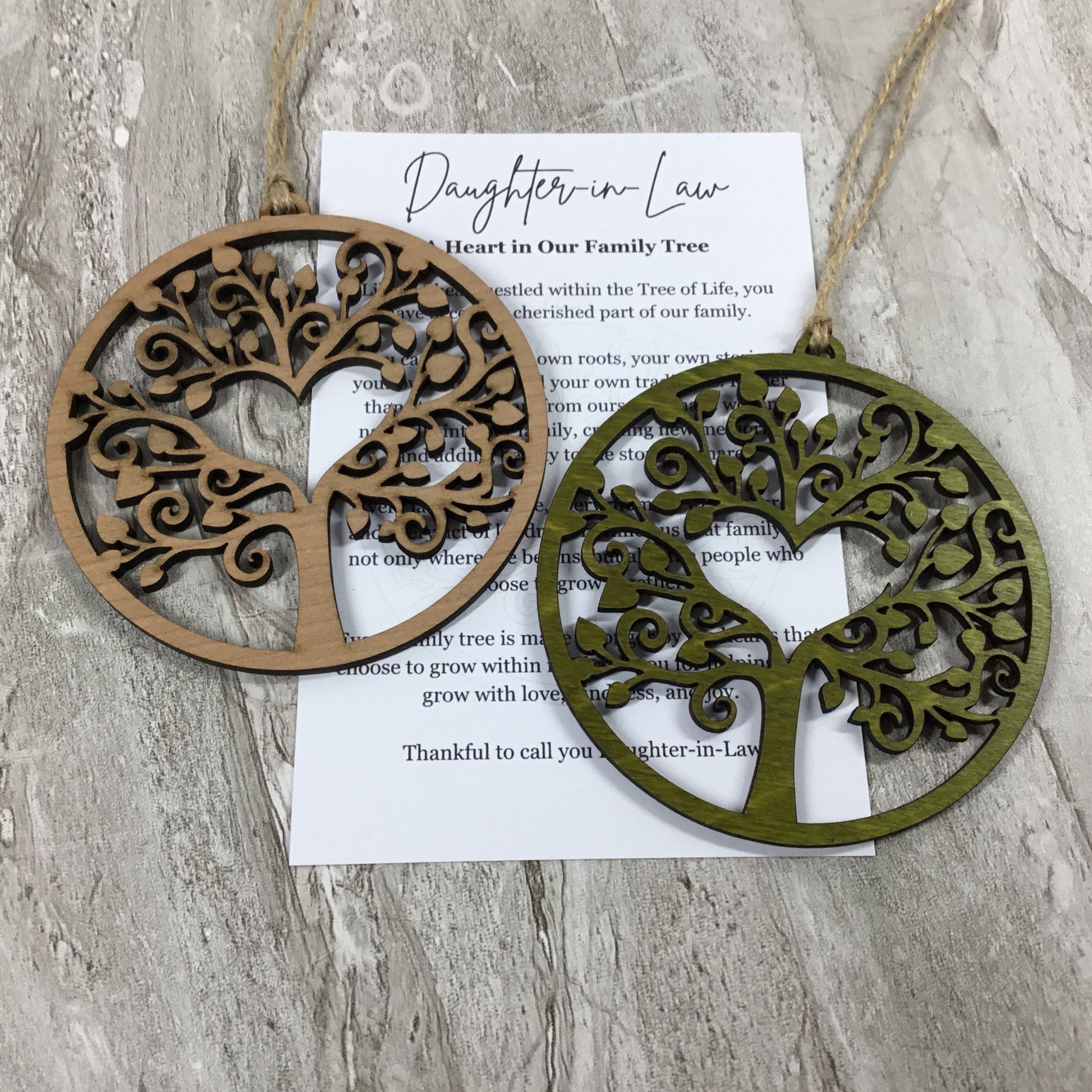 Two circular laser-cut tree of life hanging ornaments in natural wood and olive green wood finishes, displayed on a white printed card over a gray wood-textured background.