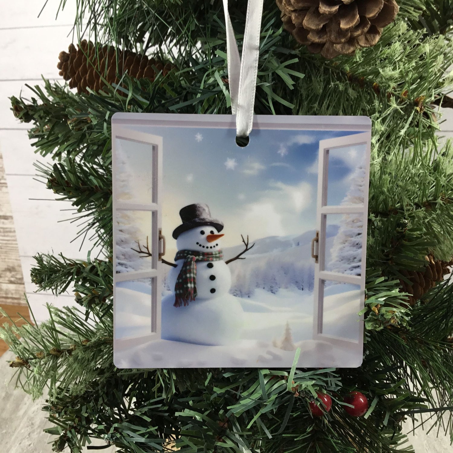 A square Christmas ornament featuring a printed winter scene of a snowman standing in front of open snowy windows, hanging from a white ribbon on a green pine tree.