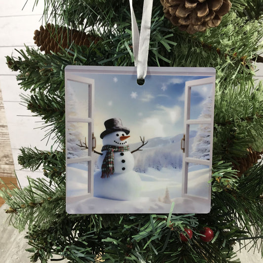 A square Christmas ornament featuring a printed winter scene of a snowman standing in front of open snowy windows, hanging from a white ribbon on a green pine tree.