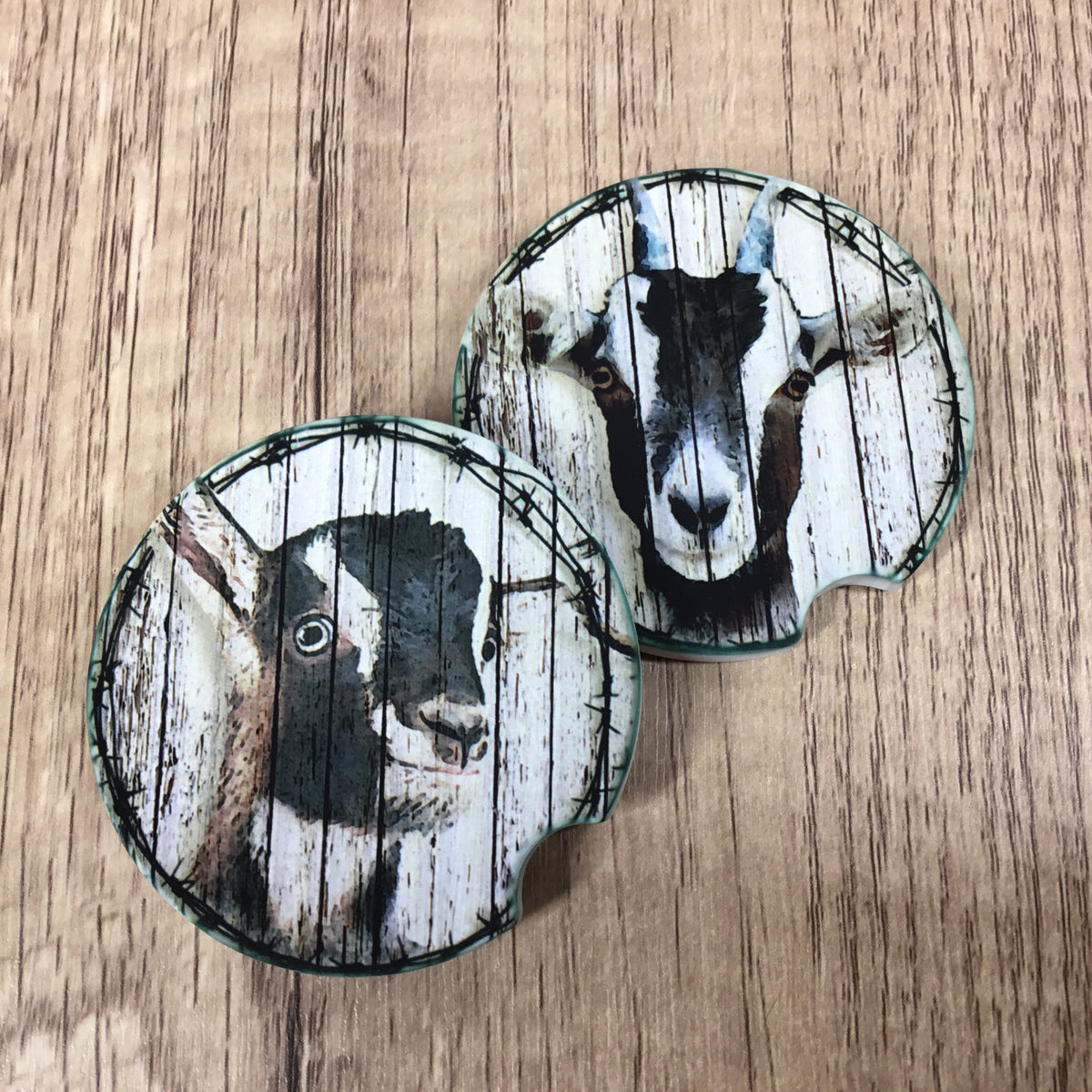 Two round car coasters featuring a rustic wood-plank design with a black and white goat portrait, displayed on a light brown wood-grain surface.