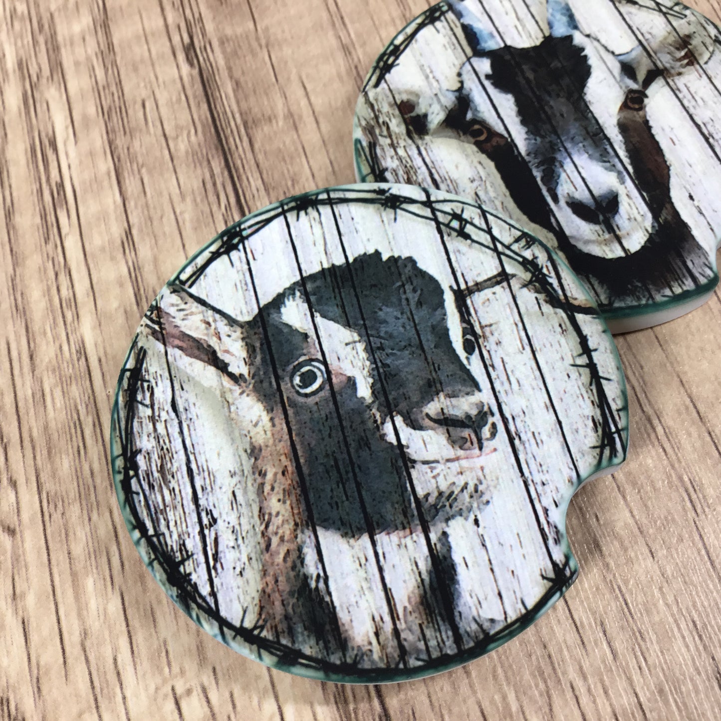 Two round coasters featuring a rustic printed design of a black and white goat face over distressed white wood planks, with a thin dark border, shown on a light brown wood-grain surface.