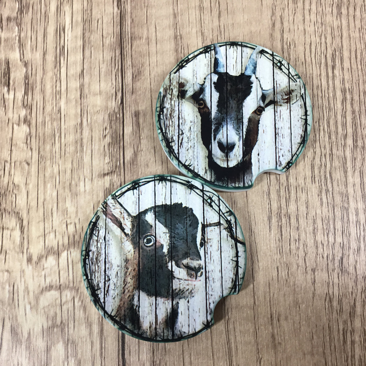 Two round coasters featuring a rustic printed goat design on a whitewashed wood plank background, displayed on a light brown wood surface.