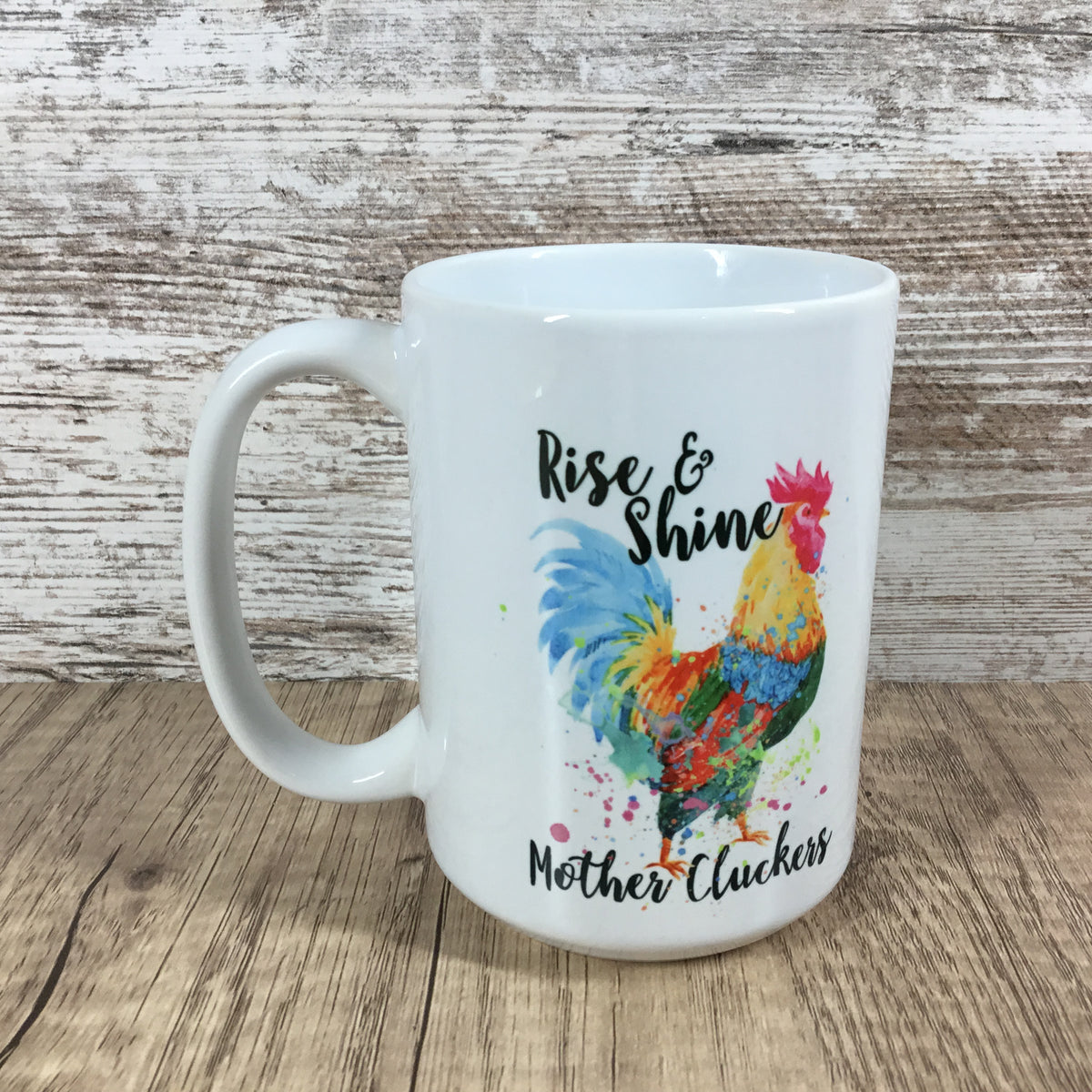 A white ceramic mug featuring a colorful watercolor-style rooster illustration and the phrases 'Rise & Shine' and 'Mother Cluckers' in black script, set against a rustic whitewashed wood background.