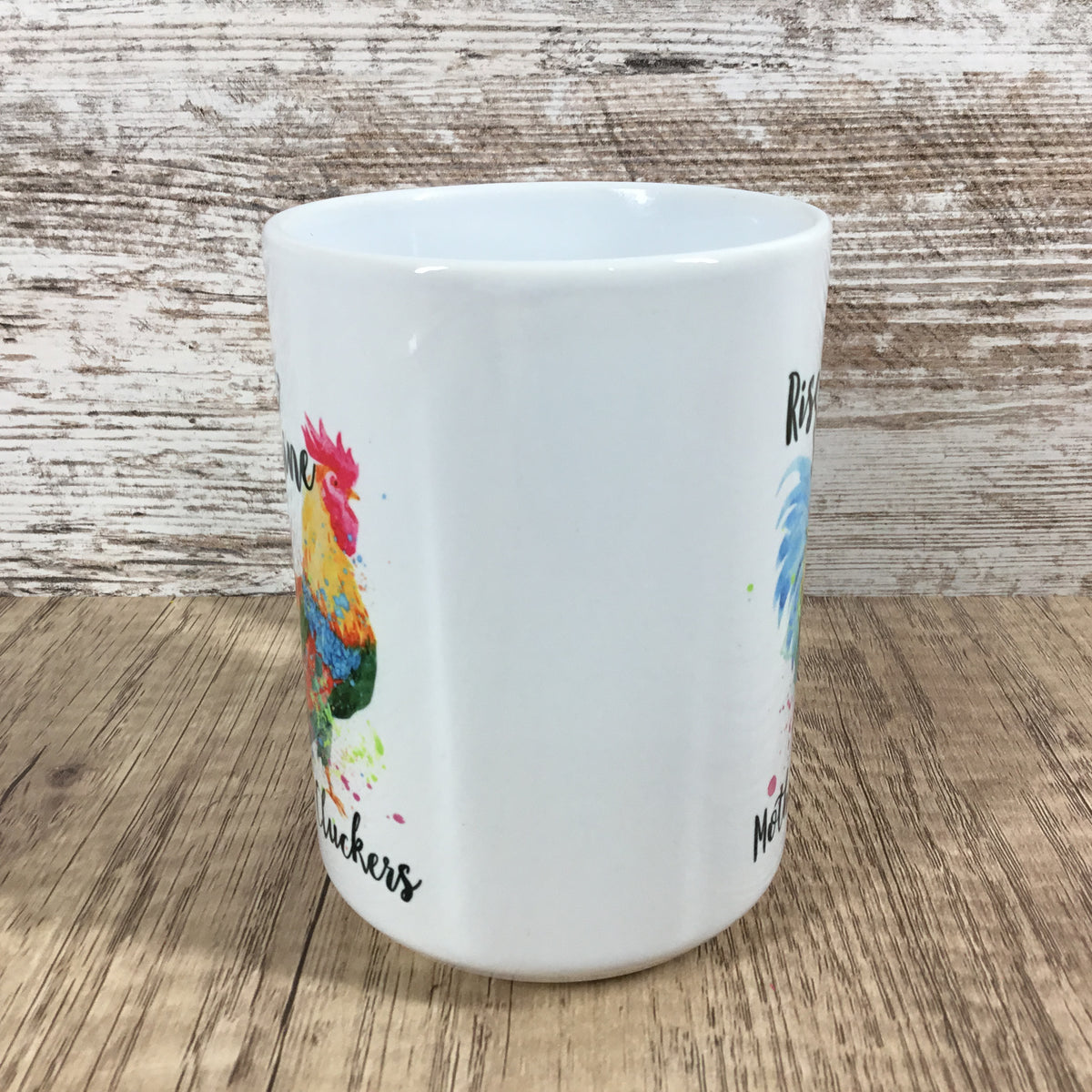 A white ceramic mug featuring colorful watercolor illustrations of chickens and black script text, set against a rustic wood background.