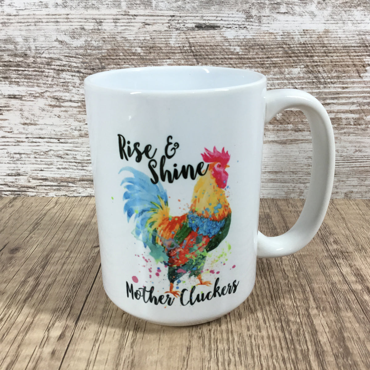 A white ceramic coffee mug with a colorful watercolor rooster design and the text 'Rise & Shine' and 'Mother Cluckers' on a rustic wood background.