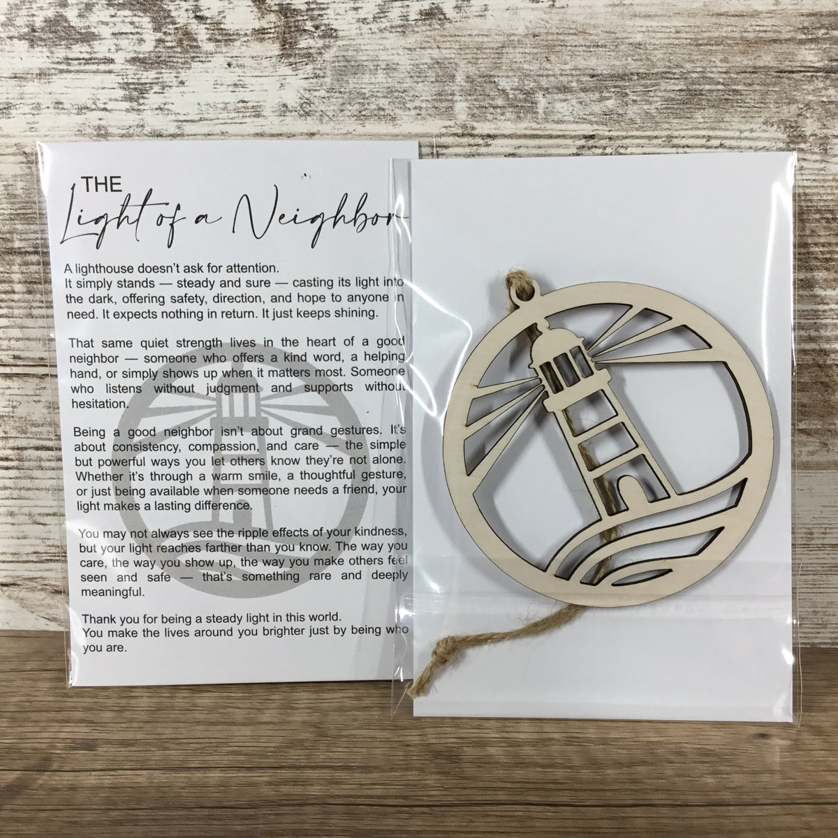 A packaged wooden lighthouse-shaped ornament with a printed message card titled 'The Light of a Neighbor,' placed on a rustic wood surface.