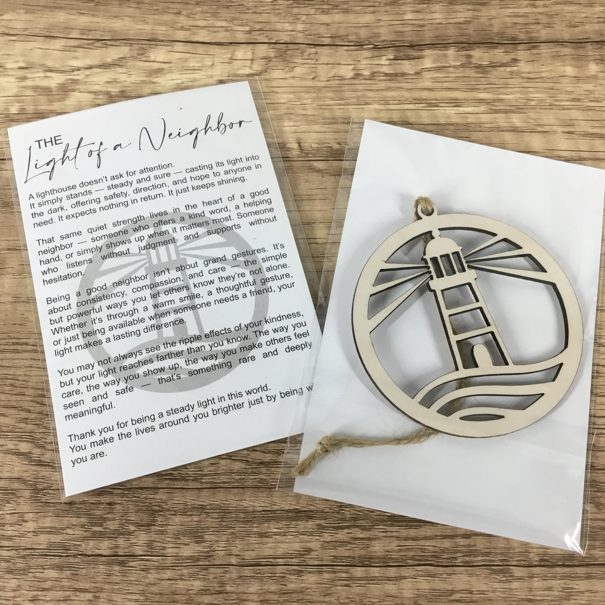 A laser-cut wooden lighthouse ornament in a circular frame, shown next to its printed message card on a white backing card, all inside a clear plastic bag on a wooden tabletop.