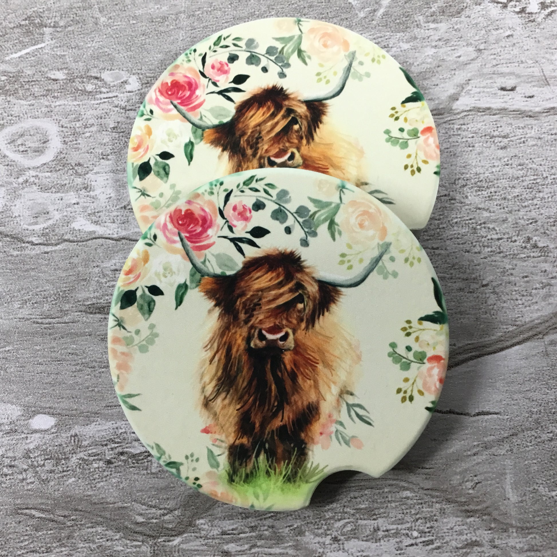 Two round car coasters with highland cow design on a floral background on a wooden surface.