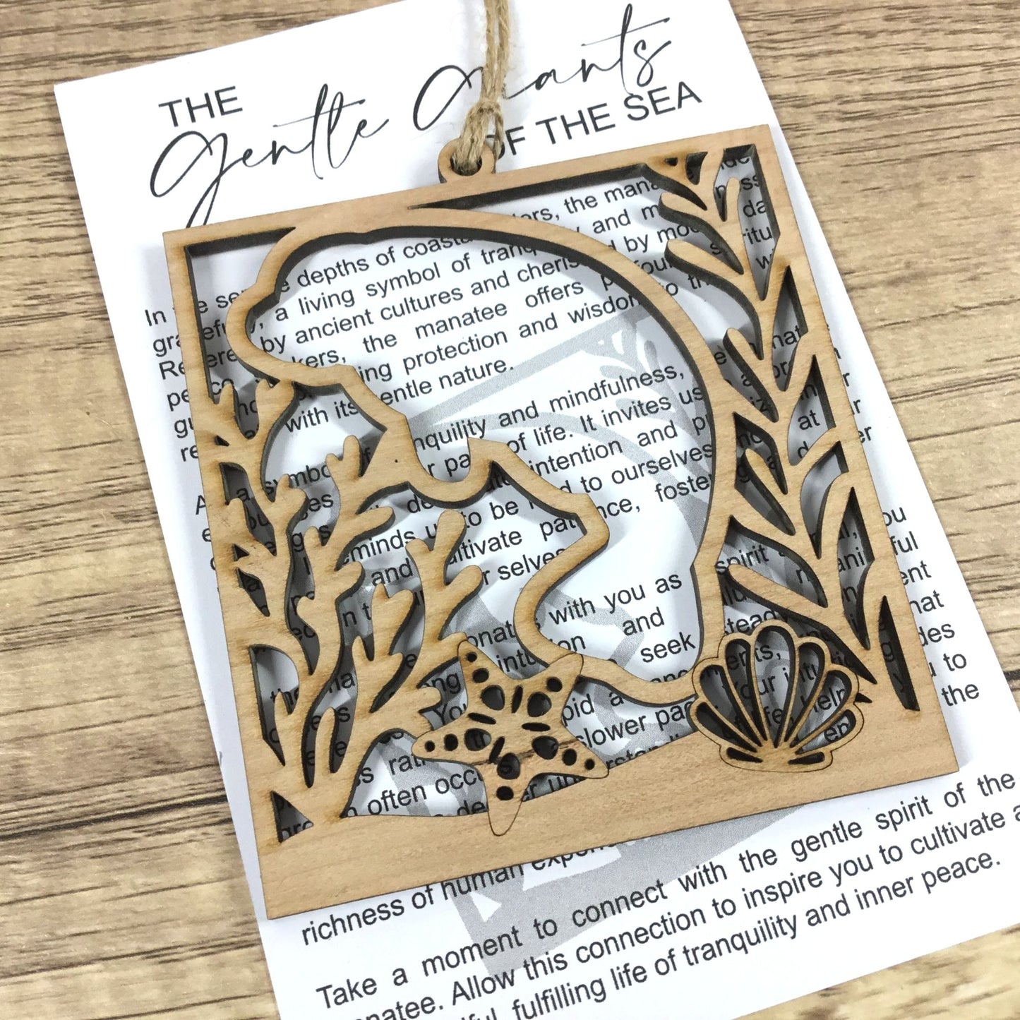 A laser-cut wooden hanging ornament featuring a manatee and seashell design over an underwater coral scene, displayed on a book page over a wooden surface.