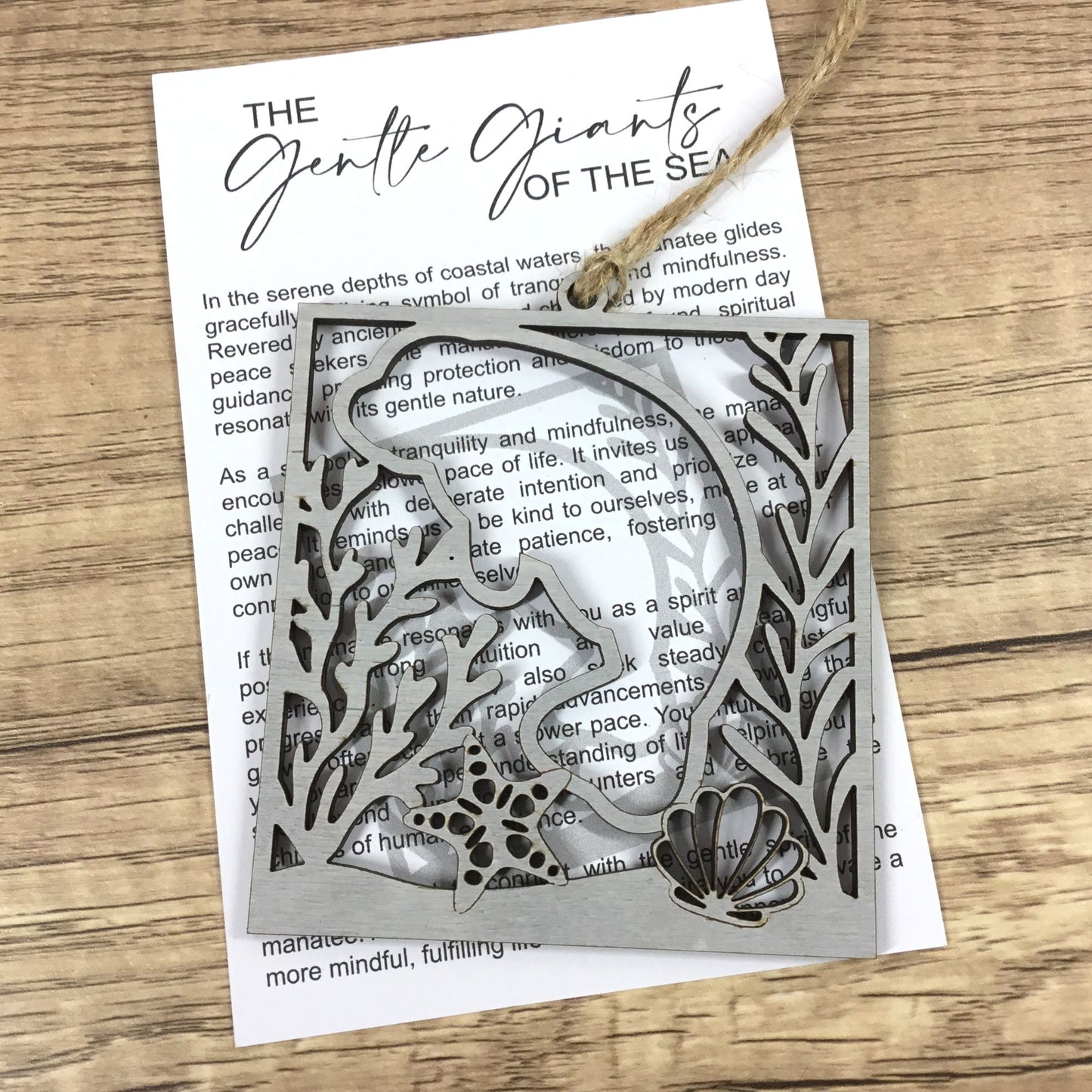 A laser-cut manatee ornament featuring  displayed on top of a printed card with a twine bow, set on a wooden surface.