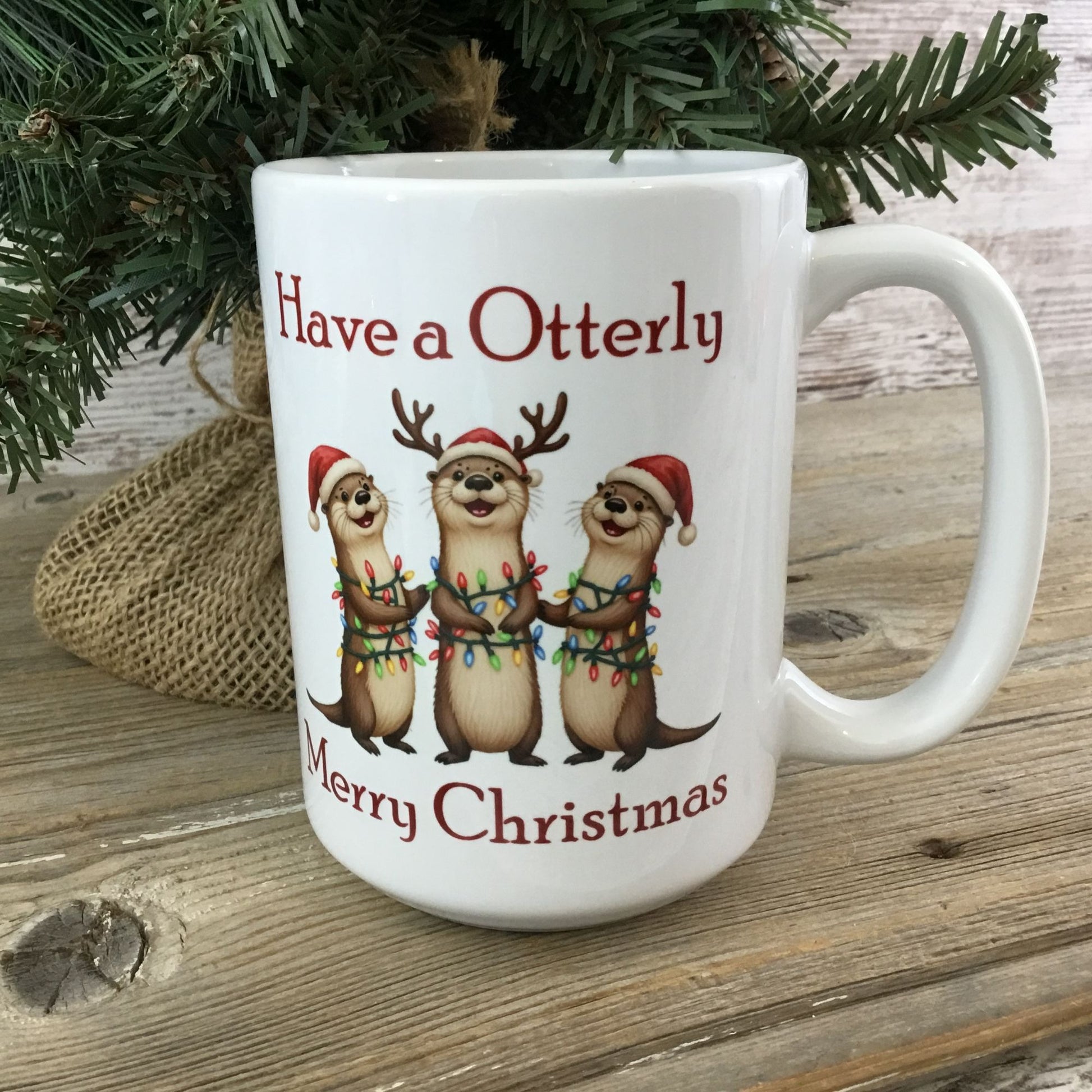 White mug with Christmas-themed design featuring otters and text, placed on a wooden surface with a festive background.
