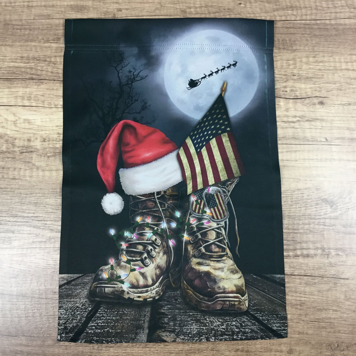 A rectangular garden flag laid flat on a light wood surface. The flag features a printed holiday scene with camouflage combat boots, a red Santa hat, an American flag, colorful string lights, and Santa's sleigh against a full moon on a dark night sky.