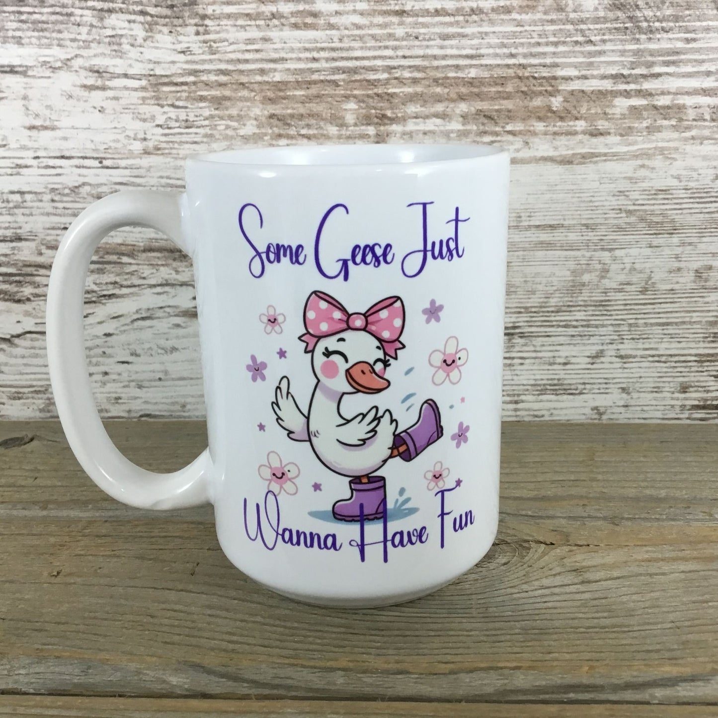 White mug with a cartoon goose wearing a pink bow and purple rain boots, text 'Some Geese Just Wanna Have Fun' on a wooden surface.
