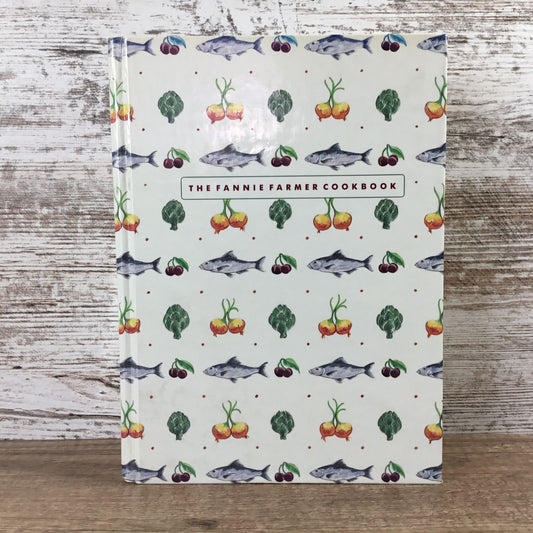 Cookbook titled 'The Fannie Farmer Cookbook' with vegetable and fish pattern on a wooden surface