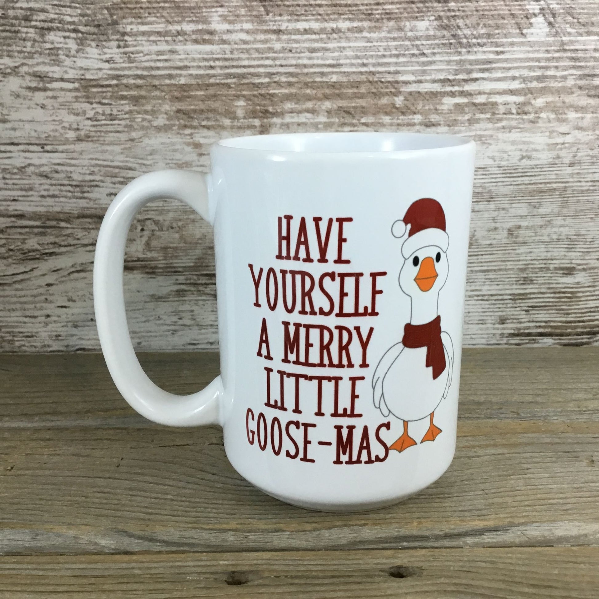White mug with a cartoon goose wearing a Santa hat and scarf, text 'Have Yourself a Merry Little Goose-Mas' on a wooden surface.