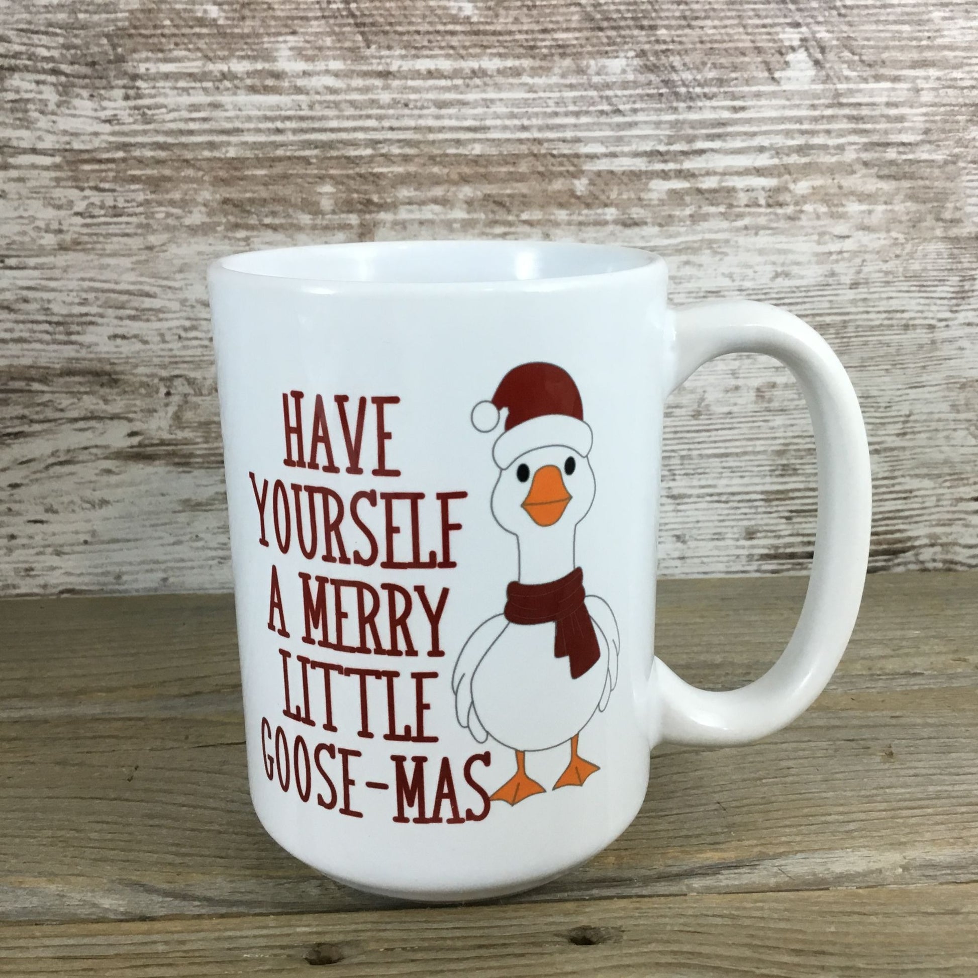 White mug with a cartoon goose wearing a Santa hat and scarf, and text 'Have Yourself a Merry Little Goose-Mas' on a wooden surface.