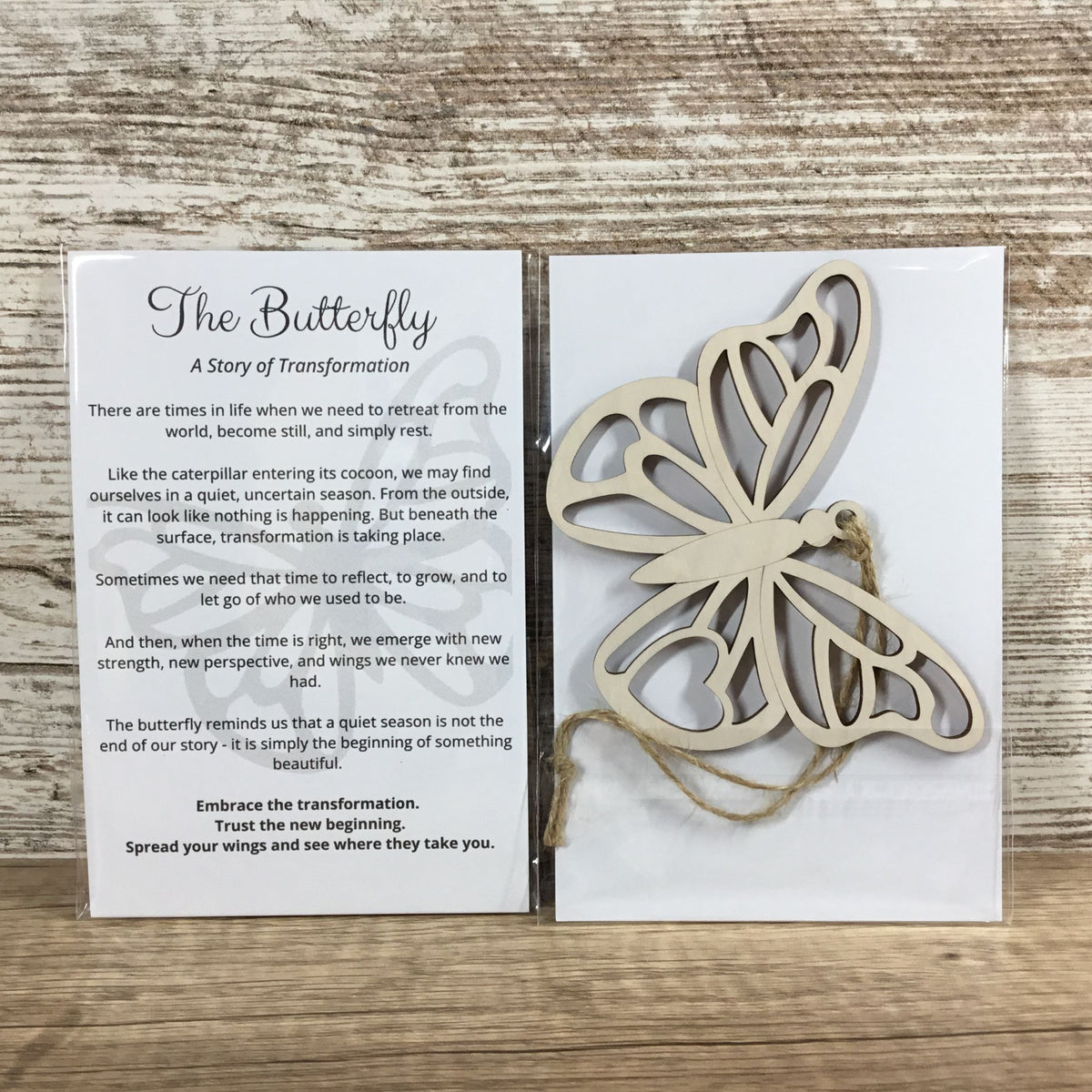 A wooden butterfly-shaped ornament displayed next to a printed card titled 'The Butterfly: A Story of Transformation' on a light wood background.