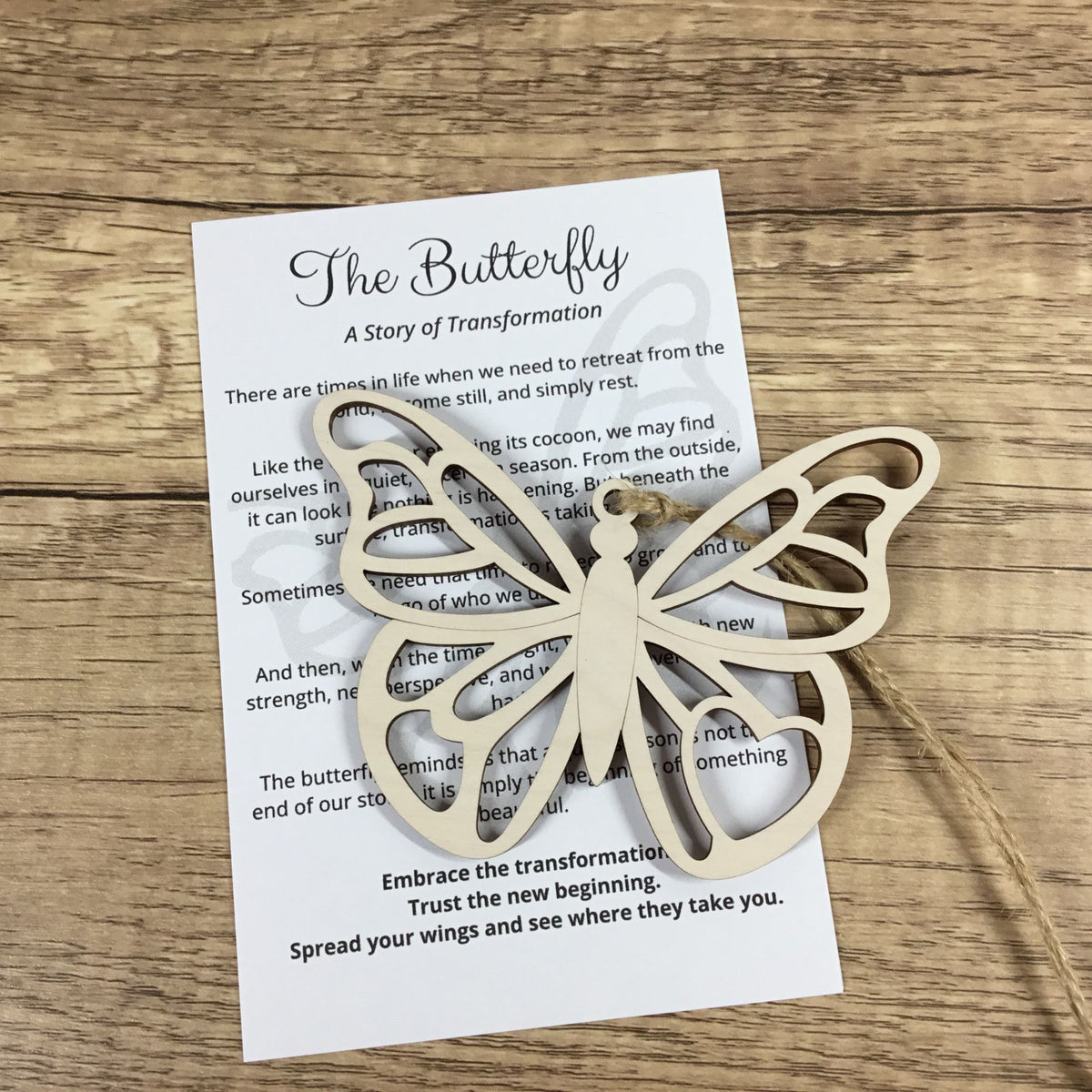 A light natural wood butterfly-shaped ornament or keyring is displayed on top of a printed card titled 'The Butterfly: A Story of Transformation' on a wooden tabletop.